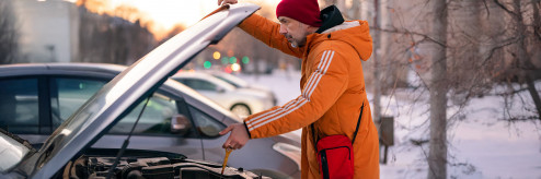 Mężczyzna w pomarańczowej kurtce i czerwonej czapce sprawdza olej silnikowy w samochodzie z otwartą maską podczas zimowego poranka.