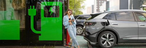 Kobieta w jasnym ubraniu oparta o zieloną stację ładowania, ładująca srebrny samochód elektryczny na tle miejskiego krajobrazu