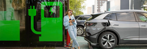 Kobieta w jasnym ubraniu oparta o zieloną stację ładowania, ładująca srebrny samochód elektryczny na tle miejskiego krajobrazu