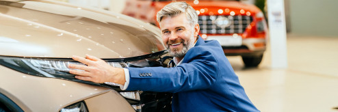 Uśmiechnięty mężczyzna w garniturze obejmuje nowoczesny samochód w salonie, symbolizując radość i emocjonalne przywiązanie do idealnie dopasowanego auta.