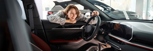 Uśmiechnięta kobieta z entuzjazmem zagląda do wnętrza nowoczesnego samochodu w salonie – symbol swobody wyboru i radości z jazdy dzięki elastycznemu wynajmowi auta z SIXT DriveSmart.
