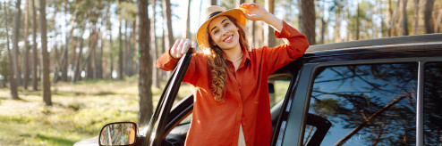 Uśmiechnięta kobieta w kapeluszu stojąca przy otwartych drzwiach czarnego samochodu na tle słonecznego, sosnowego lasu