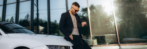 Mężczyzna w okularach przeciwsłonecznych oparty o biały samochód, patrzący na zegarek