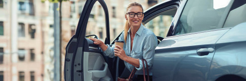 Kobieta wysiadająca z nowoczesnego samochodu z kubkiem kawy – symbol komfortowej podróży elektrykiem.