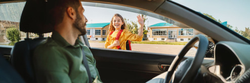 Uśmiechnięta dziewczynka w żółtej bluzie macha do mężczyzny siedzącego za kierownicą samochodu , w tle widać budynek szkolny.
