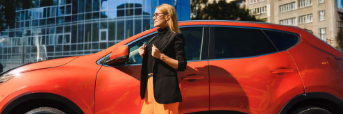 Stylowa blondynka w okularach i czarnej marynarce na tle nowoczesnych budynków, oparta o pomarańczowy SUV.