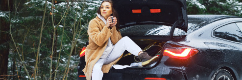 Kobieta siedząca w otwartym bagażniku samochodu w zimowym lesie, trzymająca kubek z napojem – wynajem auta na zimę SIXT DriveSmart.